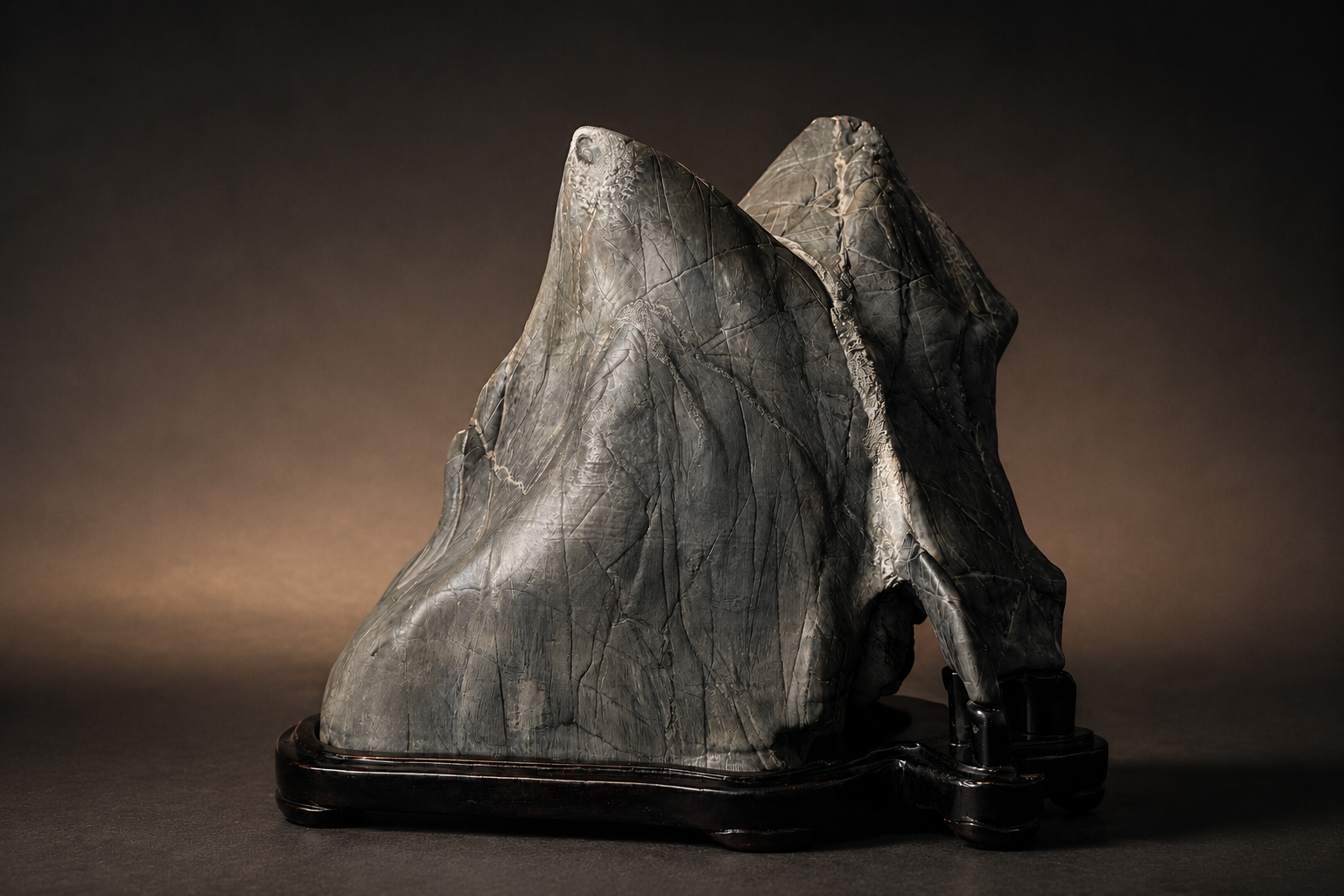 Suiseki de dos picos entrelazados, con reflejos de luz sobre la piedra, en daiza de madera oscura · colección de Marcial Yuste Blasco