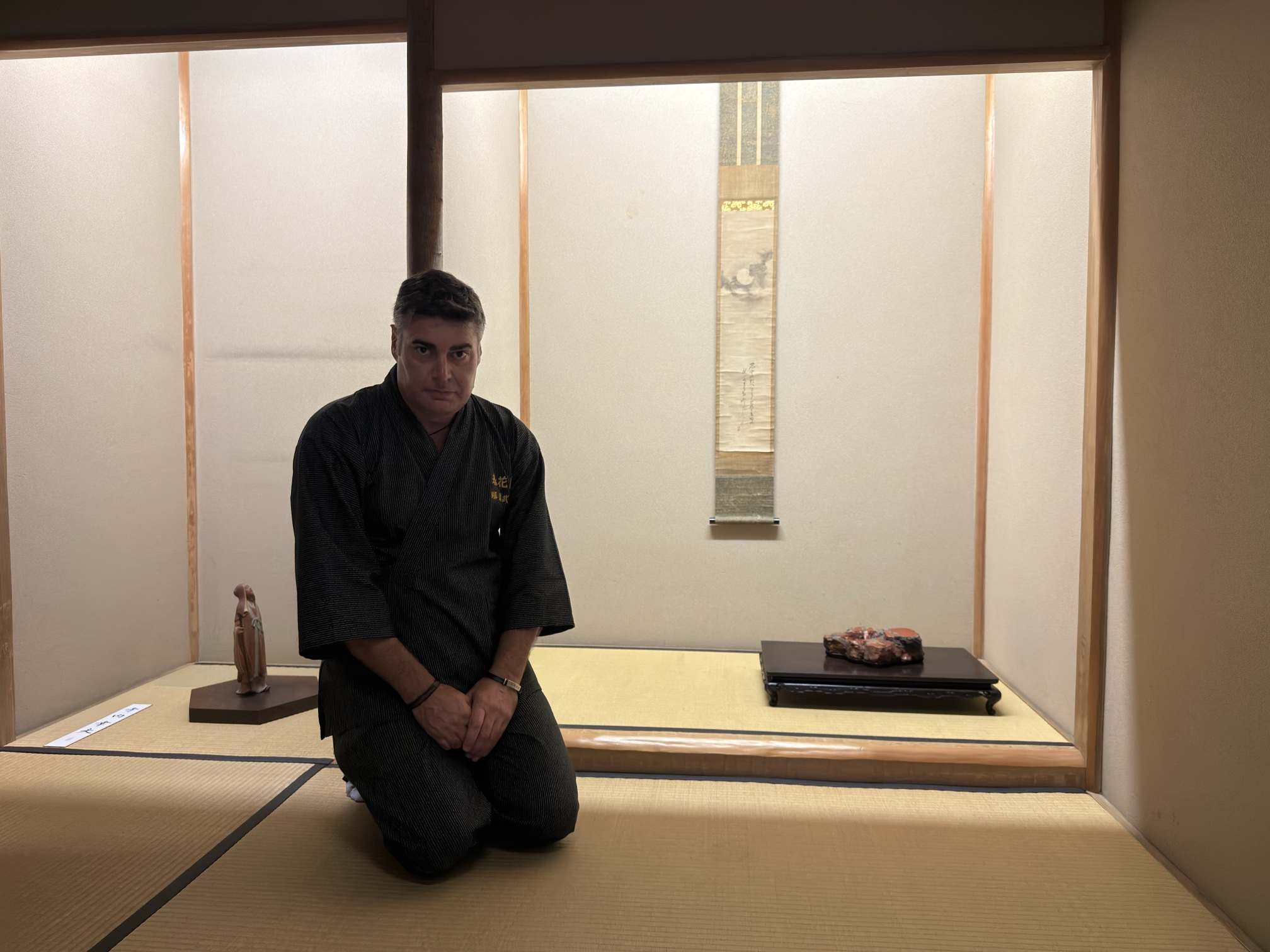 Masaru Ken, arrodillado en seiza ante el tokonoma de una sala de té, con un kakejiku, un suiseki y una pequeña figura