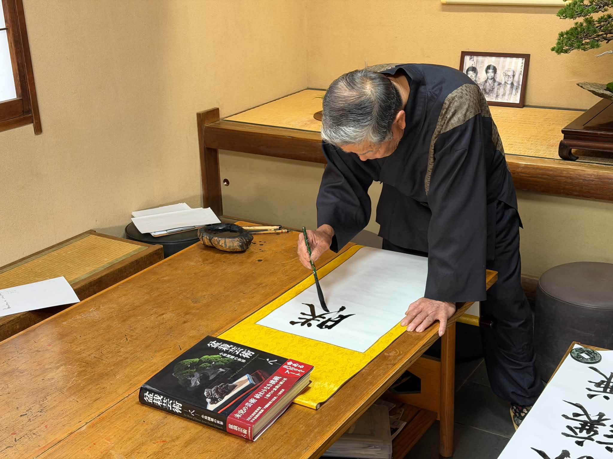 El III Iemoto Kobayashi Ippū escribe a pincel el kakejiku con el nombre artístico 勝讓謙, sobre una mesa de madera con tinta y pinceles